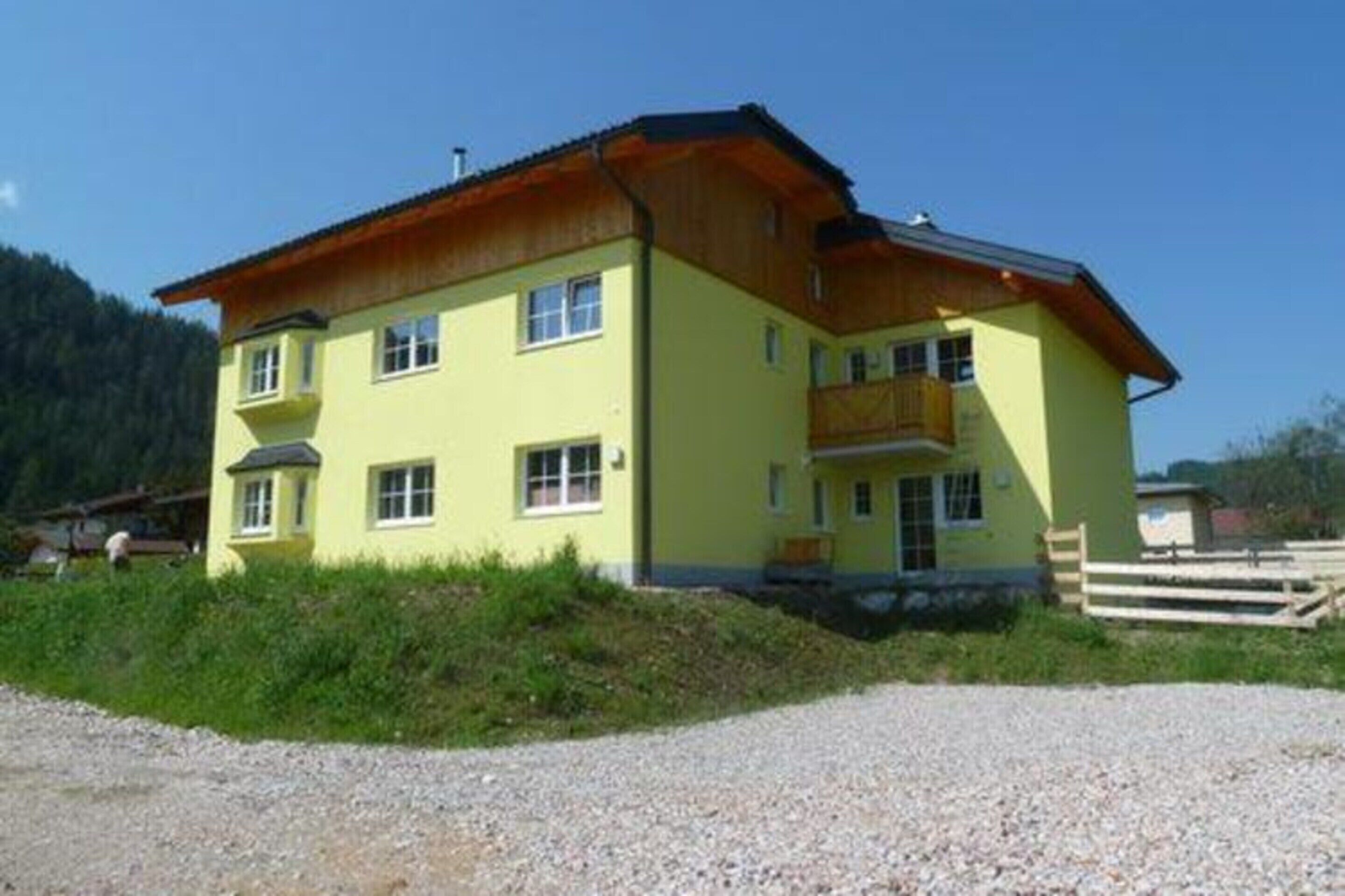 A two-story yellow building with a wooden balcony, surrounded by gravel and greenery.