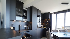 A modern kitchen with dark cabinetry, a central island, and a dining area with chairs. - Wohnung in der Nordseeregion in Strandnhhe (Wyk auf Foehr)