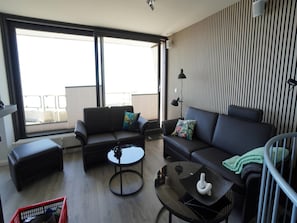 A modern living room with a sofa, a coffee table, and a small side table. There is a window providing a view of the outside. - Wohnung in der Nordseeregion in Strandnahe (Wyk auf Foehr)