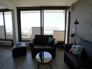 Una sala moderna con sofá, sillón y mesa de centro, ventanas amplias con vista al mar y una pared a rayas.