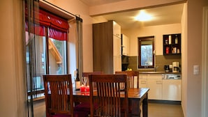 Un coin repas avec une table et des chaises en bois, une fenêtre avec des rideaux rouges et, au fond, une cuisine.