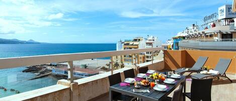A balcony with a table set for breakfast overlooking the sea, with buildings and a clear sky in the background.