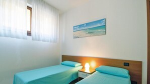 A room with two single beds, a nightstand with a lamp, a painting of a beach scene, and a window with sheer curtains. - Apartment in Villassimius Garden (Villasimius)