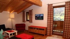 Una stanza accogliente con soffitto in legno, un divano rosso, un piccolo tavolo bianco, una panca rossa e un'ampia finestra con tende a quadri.
