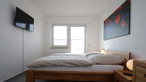 Une chambre à coucher avec un lit, une table de chevet, un téléviseur fixé au mur et une fenêtre.