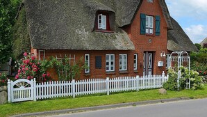 A traditional thatched-roof house with a white picket fence, brick walls, and a small garden with pink flowers. - Ferienwohnung in Oevenum auf Foehr (Oevenum)