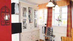 A living room with a white cabinet, a table with a board game, a yellow armchair, and a window with curtains. - Ferienwohnung in Oevenum auf Foehr (Oevenum)