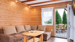 Ein gemütlicher Raum mit Holzwänden und -decke, einem braunen Sofa, einem Holztisch und einer Schiebetür aus Glas zu einem Außenbereich mit einer Holzdielenveranda und Stühlen.