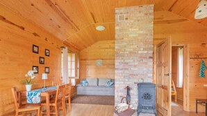 Ein gemütliches Blockhaus-Interieur mit Holzwänden, einem Kamin aus Ziegeln, einem Essbereich mit Tisch und Stühlen sowie einer Sitzecke mit Sofa.
