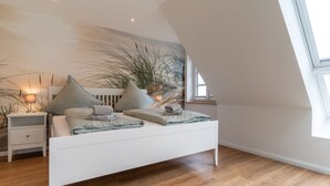 A modern bedroom with a white bed, wooden flooring, and a large window. - Doppelhaushalfte in Karolinenkoog (Karolinenkoog)