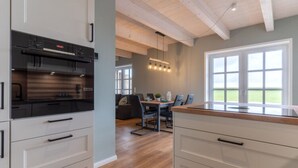 A modern kitchen with white cabinetry and a built-in oven. There is a dining area with a table and chairs, and a large window providing a view of the outdoors. - Doppelhaushalfte in Karolinenkoog (Karolinenkoog)