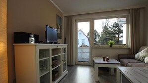 Olohuoneessa on televisiotaulu, sohva, pieni pöytä ja ikkuna, josta avautuu näkymä taloon ja puihin.