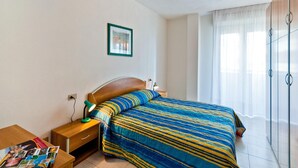 A bedroom with a bed, a bedside table with a lamp, a framed picture on the wall, and a window with curtains. - Residence Cristoforo Colombe Caorle (Caorle)