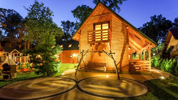 A wooden cabin with a porch and a swing set, surrounded by trees and a paved area. - Chalet in Pobierowo Near Baltic Sea Beach (Rewal)