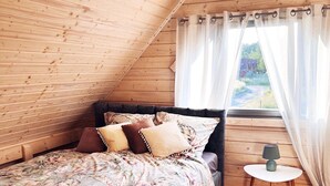 A cozy bedroom with a sloped ceiling, a bed with floral bedding, a bedside table with a lamp, and a window with sheer curtains. - Holiday Home in Niechorze With Garden (Rewal)