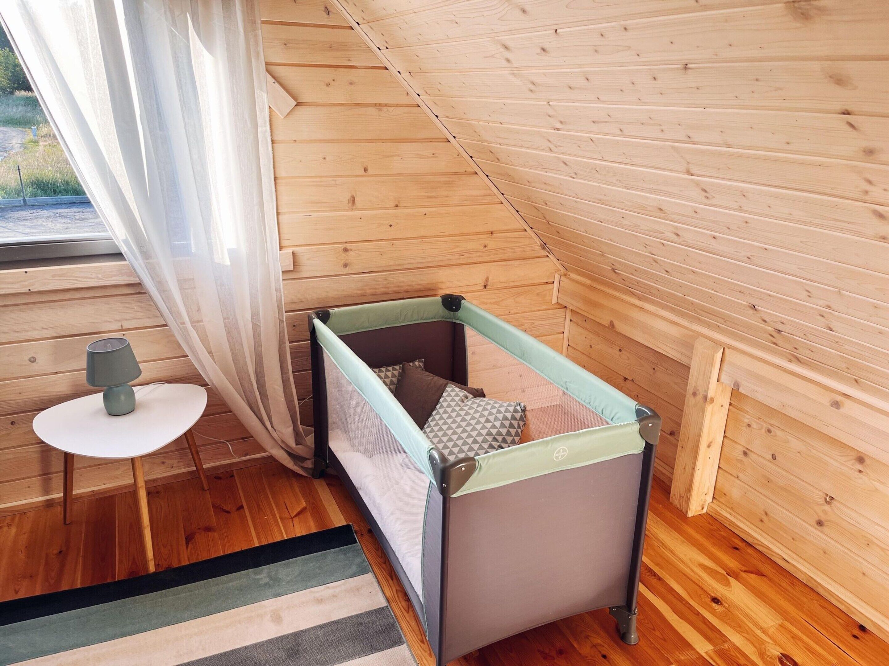 A cozy room with a wooden sloped ceiling, a small bed with a checkered pillow, a round table with a lamp, and a view of greenery outside.