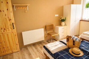 Une chambre avec une porte en bois, un radiateur, une chaise, un lit recouvert d’une couverture bleue, un panier et une fenêtre donnant sur un paysage agréable.