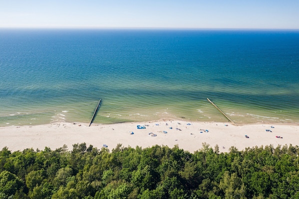 Vue aérienne d’une plage aux eaux d’un bleu limpide, bordée d’un rivage sablonneux, avec une zone boisée au premier plan.