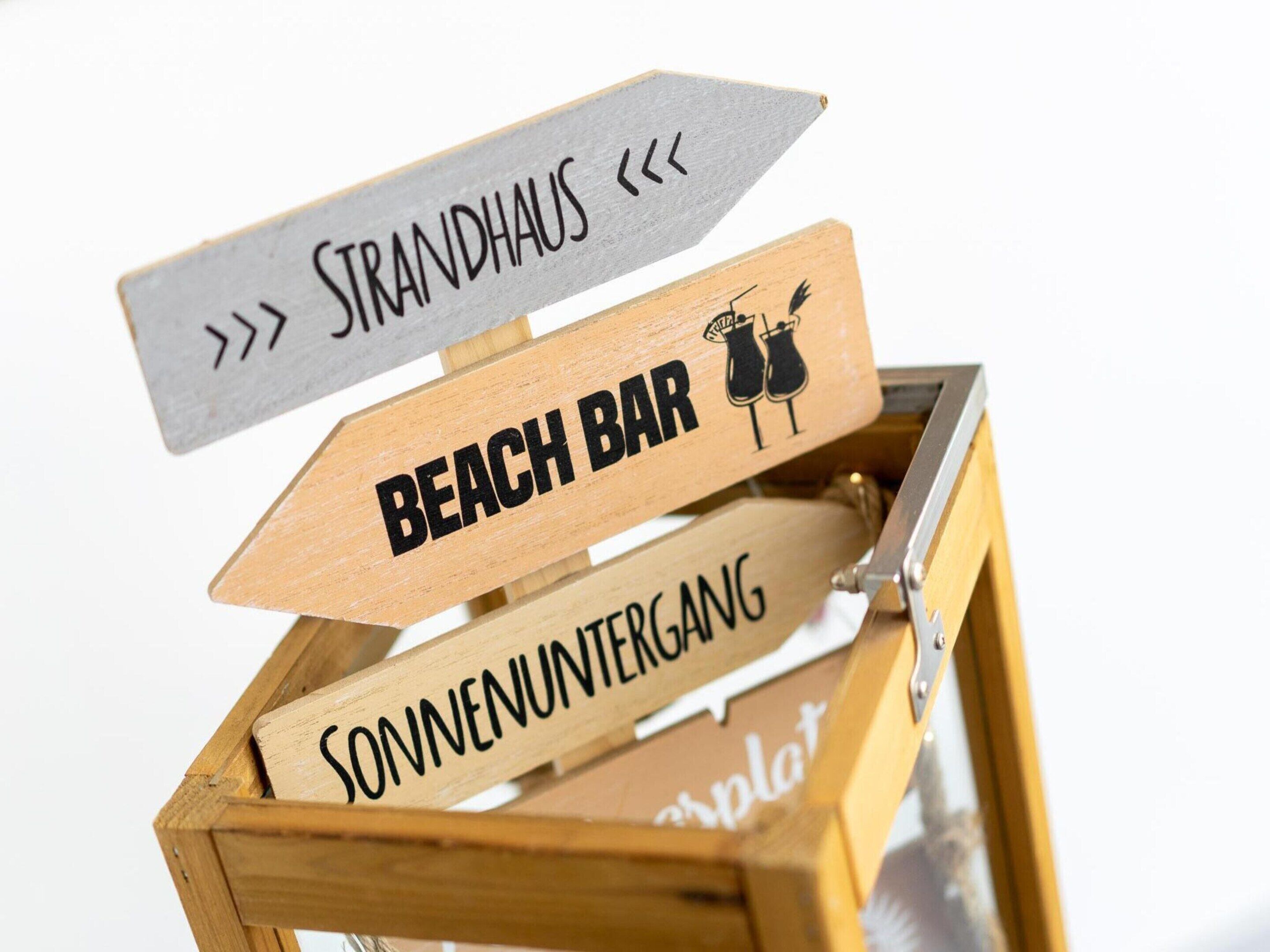 A wooden box with directional signs indicating 'Strandhaus', 'Beach Bar', and 'Sonnenuntergang'.