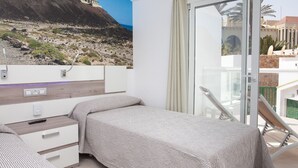 A bedroom with a bed, a bedside table, a small drawer unit, and a large window with a view of a rocky landscape and a building. - TAO Caleta Playa, Corralejo (La Oliva)