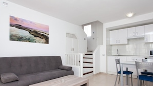 A modern living room with a grey sofa, a wooden coffee table, and a kitchen area with white cabinets and a dining set. - TAO Caleta Playa, Corralejo (La Oliva)
