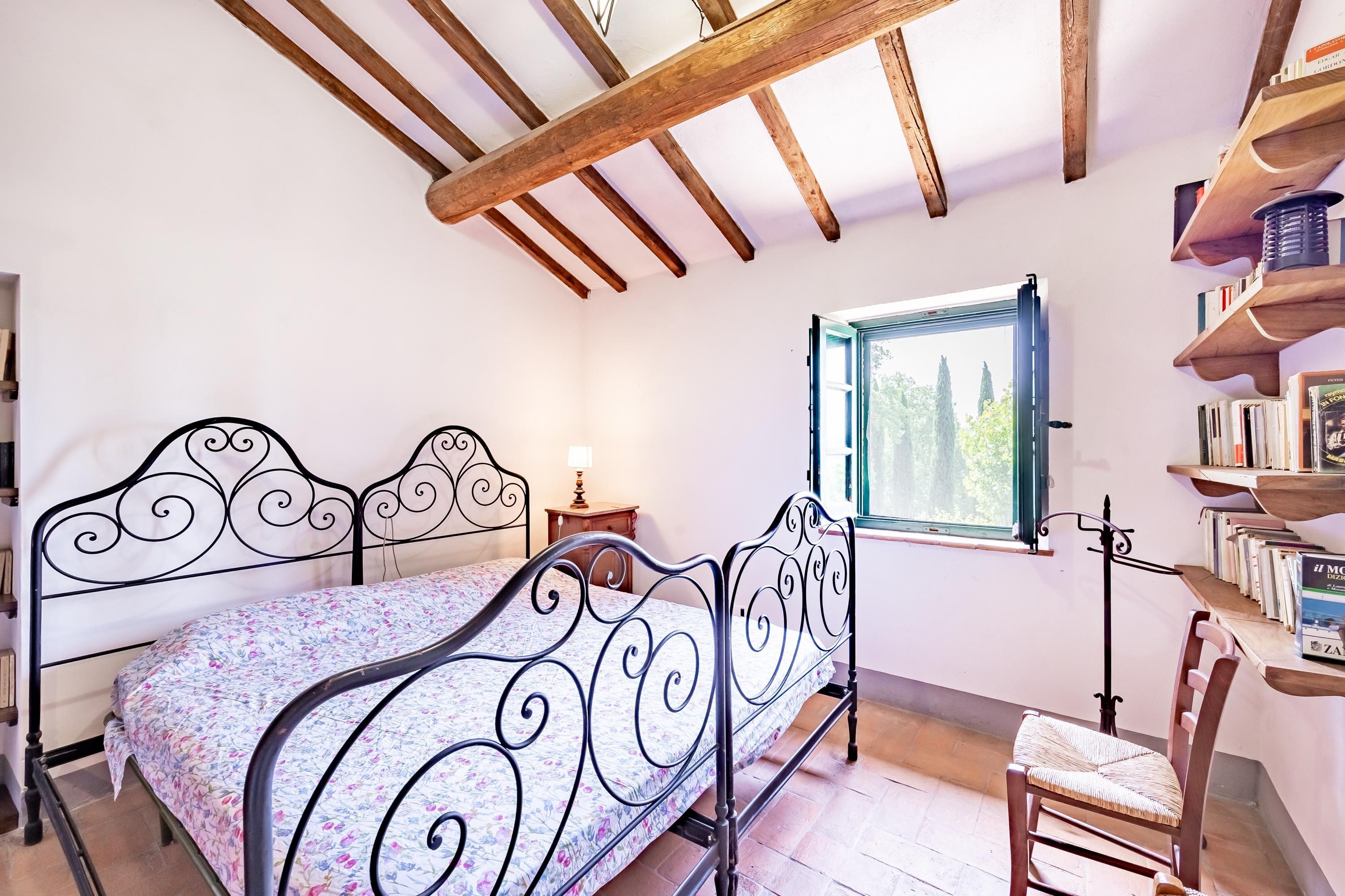 A bedroom with a metal bed frame, a window with a view of trees, and a wooden bookshelf.