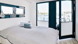 A bedroom with a large bed, a window with a sailboat model, and a view of a marina through an open door. - Houseboat in Egernsund at the Marina Minde (Broager)