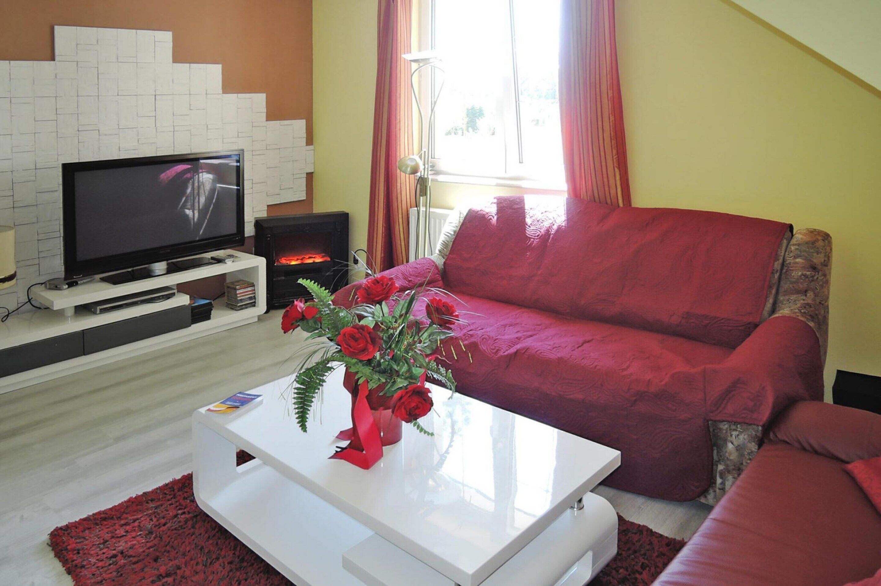 A living room with a flat-screen TV, a red sofa, a white coffee table with a vase of red roses, and a fireplace.