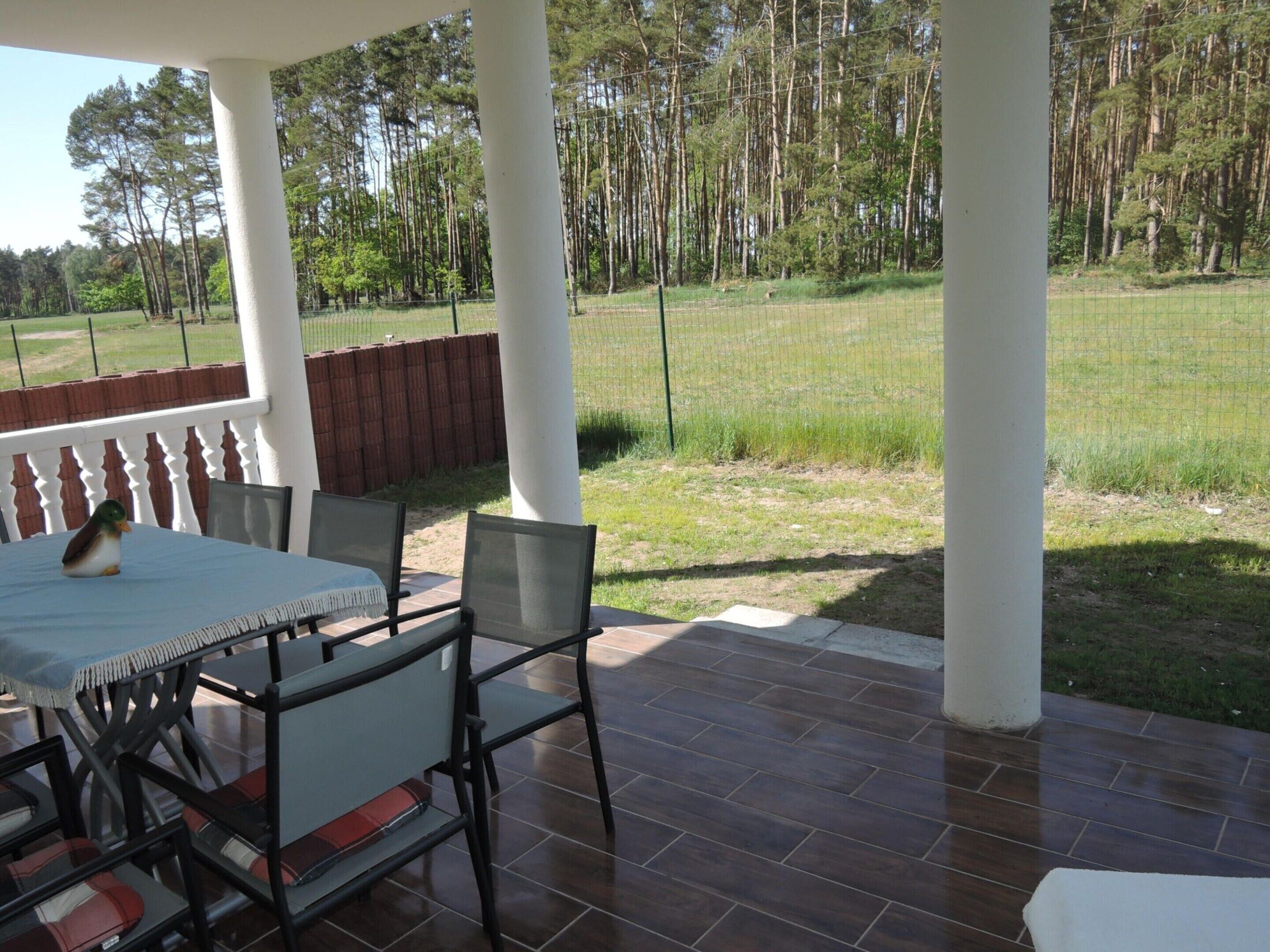 An outdoor patio with a table and chairs, surrounded by a white fence and tall trees in the background.
