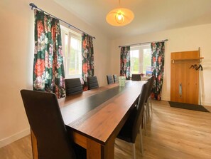 A dining area with a long wooden table, black chairs, floral curtains, and a wooden wardrobe with hanging items. - Holiday Home in Nowe Warpno Near Harbor (Nowe Warpno)