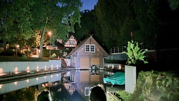 A wooden house with a reflection in a calm water body, surrounded by trees and a well-lit pathway.