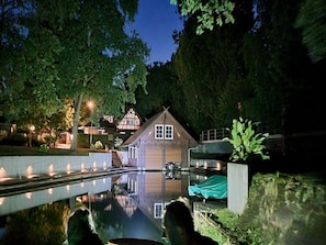 A wooden house with a reflection in a calm water body, surrounded by trees and a well-lit pathway.