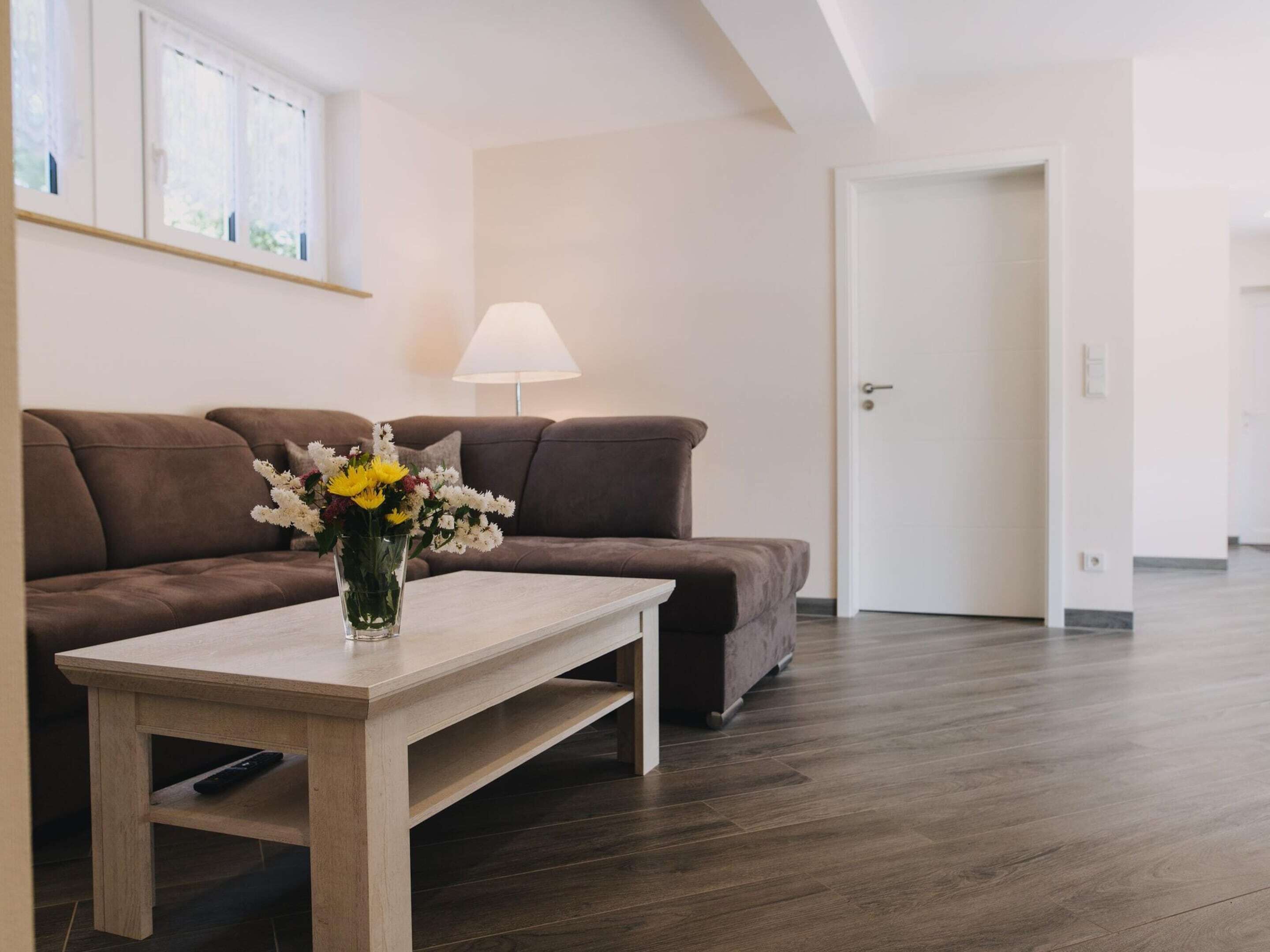 A modern living room with a brown sofa, a wooden coffee table with a vase of flowers, a lamp, and a door in the background.