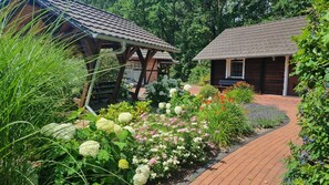 Un sendero en un jardín conduce a una cabaña de madera con techo de tejas, rodeada de vegetación exuberante y plantas con flores.