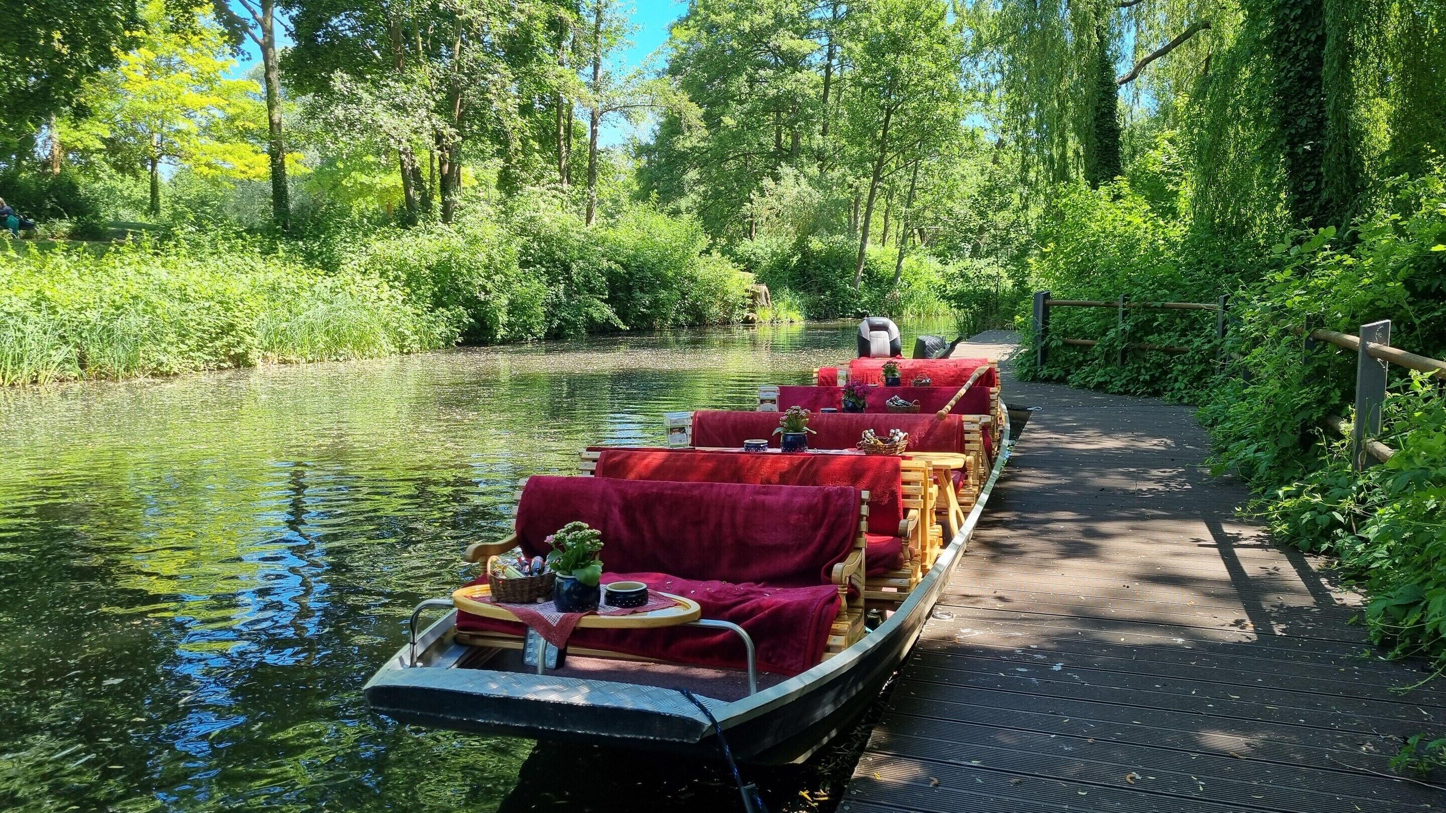 Un barco fluvial estrecho, con manteles rojos y bancos de madera, atracado en un sendero a orillas del río rodeado de vegetación.