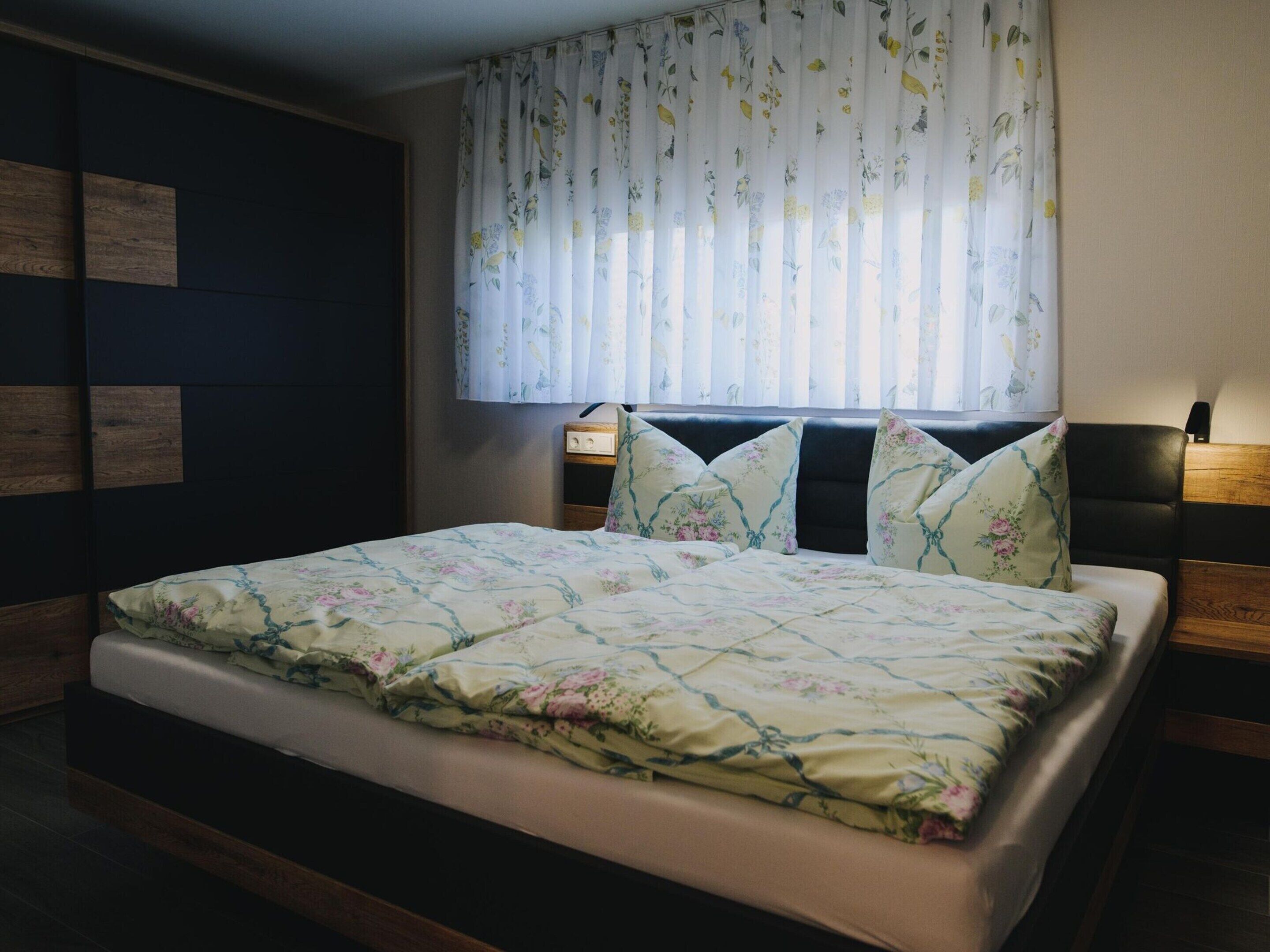 Een slaapkamer met een bed, bloemenbeddels, een nachtkastje, een lamp en een raam met bloemen gordijnen.