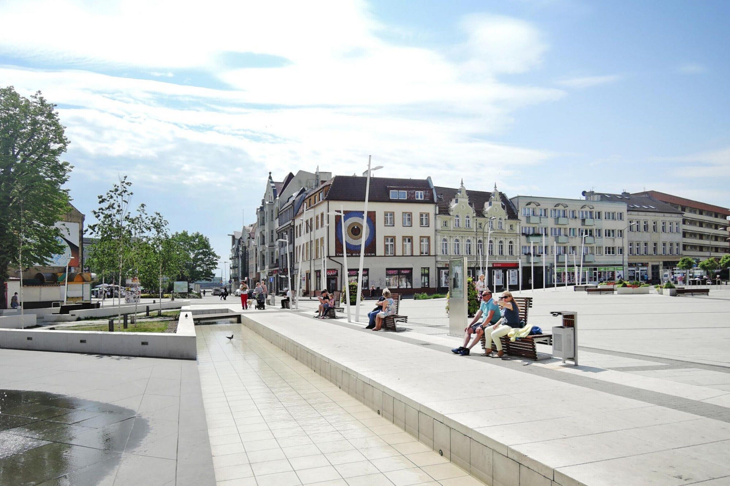 Ein öffentlicher Platz mit Bänken und einem Brunnen, umgeben von historischen Gebäuden mit unterschiedlicher Architektur.