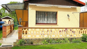 Une maison de plain-pied avec un toit rouge, une fenêtre ornée d’un rideau et un petit porche doté d’une balustrade en treillis.