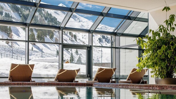 Una piscina interior con paredes de vidrio transparente, sillones de mimbre y una planta en maceta.