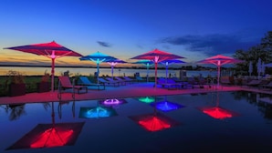 Un espace aménagé au bord de la piscine, avec plusieurs chaises longues et des parasols, une étendue d’eau calme en arrière-plan et un ciel dégagé.