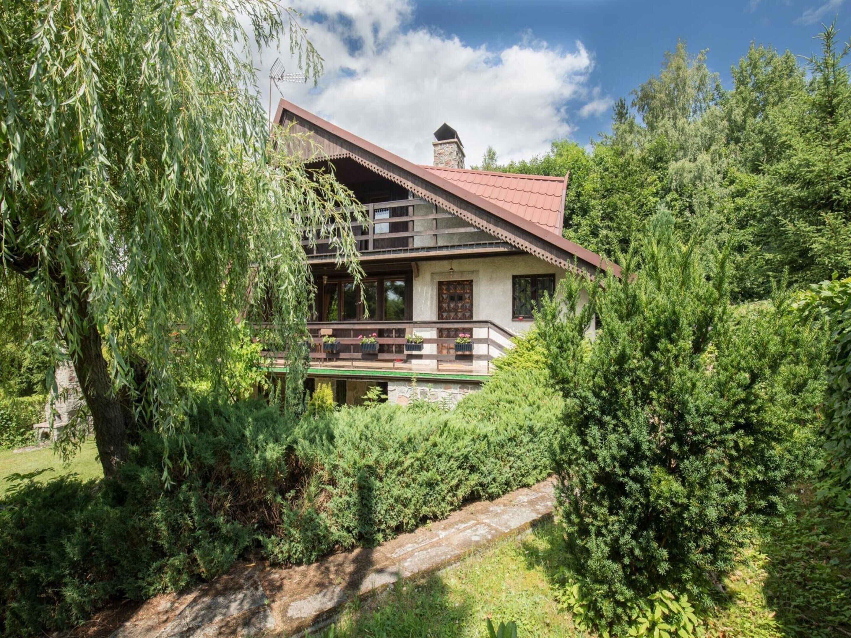 Une maison à deux étages dotée d’un balcon, entourée d’une végétation luxuriante et d’arbres.