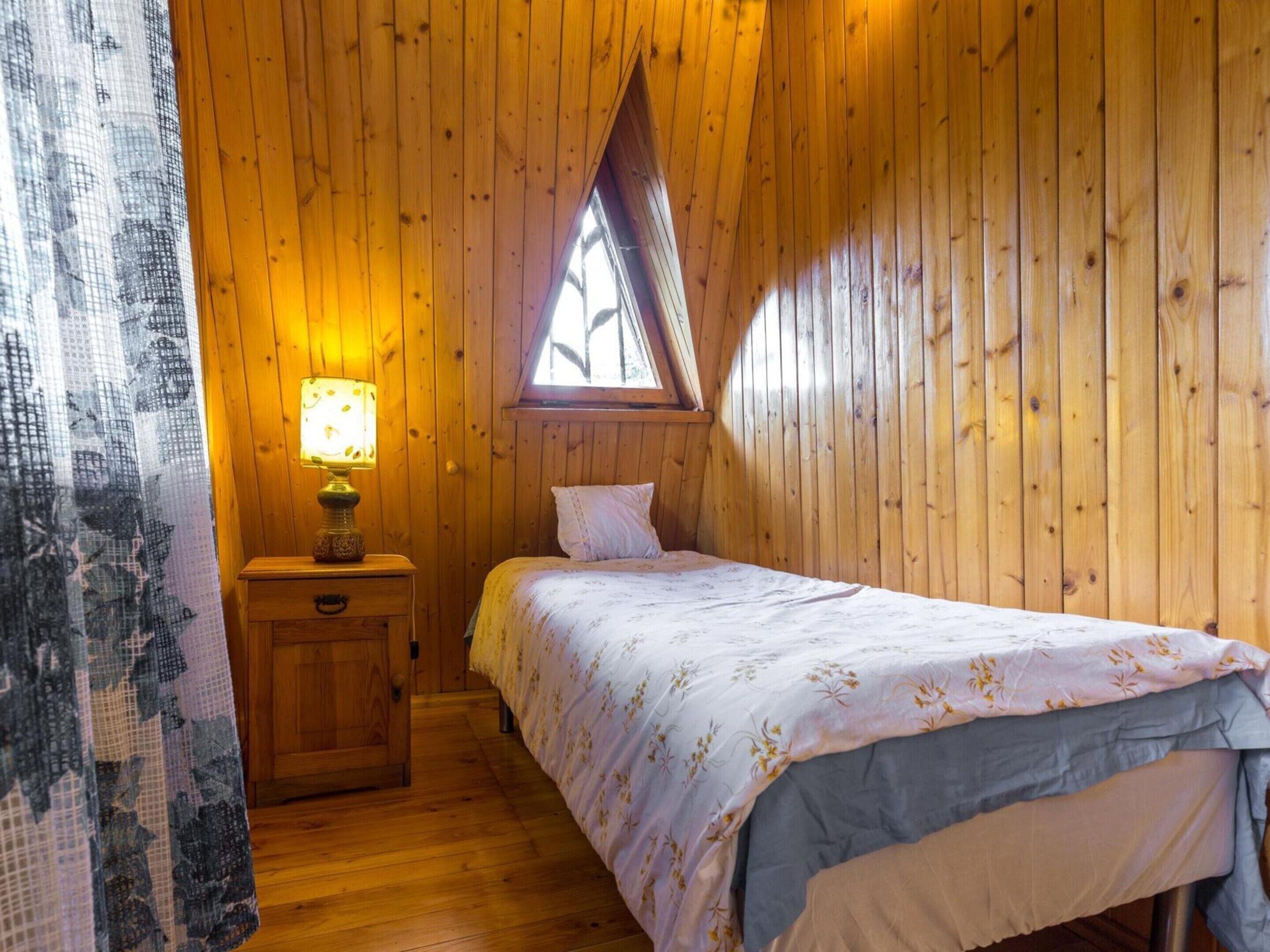 Une pièce chaleureuse aux murs en bois, avec un lit simple recouvert d’une couvre-lit à motifs floraux, une table de chevet avec une lampe et une fenêtre surmontée d’un toit triangulaire.