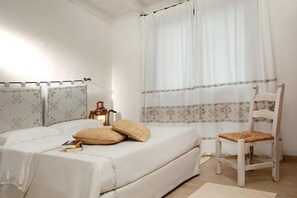 Un dormitorio con cama, almohadas, una silla, una mesita de noche con lámpara y un ventanal con cortinas.