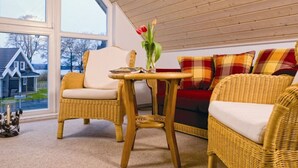 Um quarto acolhedor com móveis de vime, uma mesa com um vaso de flores e vista para uma casa através de uma grande janela.