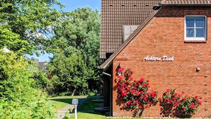 A brick building with a sign reading "Achtern Dick" and a garden with red flowers. - Ferienwohnung Direkt am Deich in St.peter-ording (St. Peter-Ording)