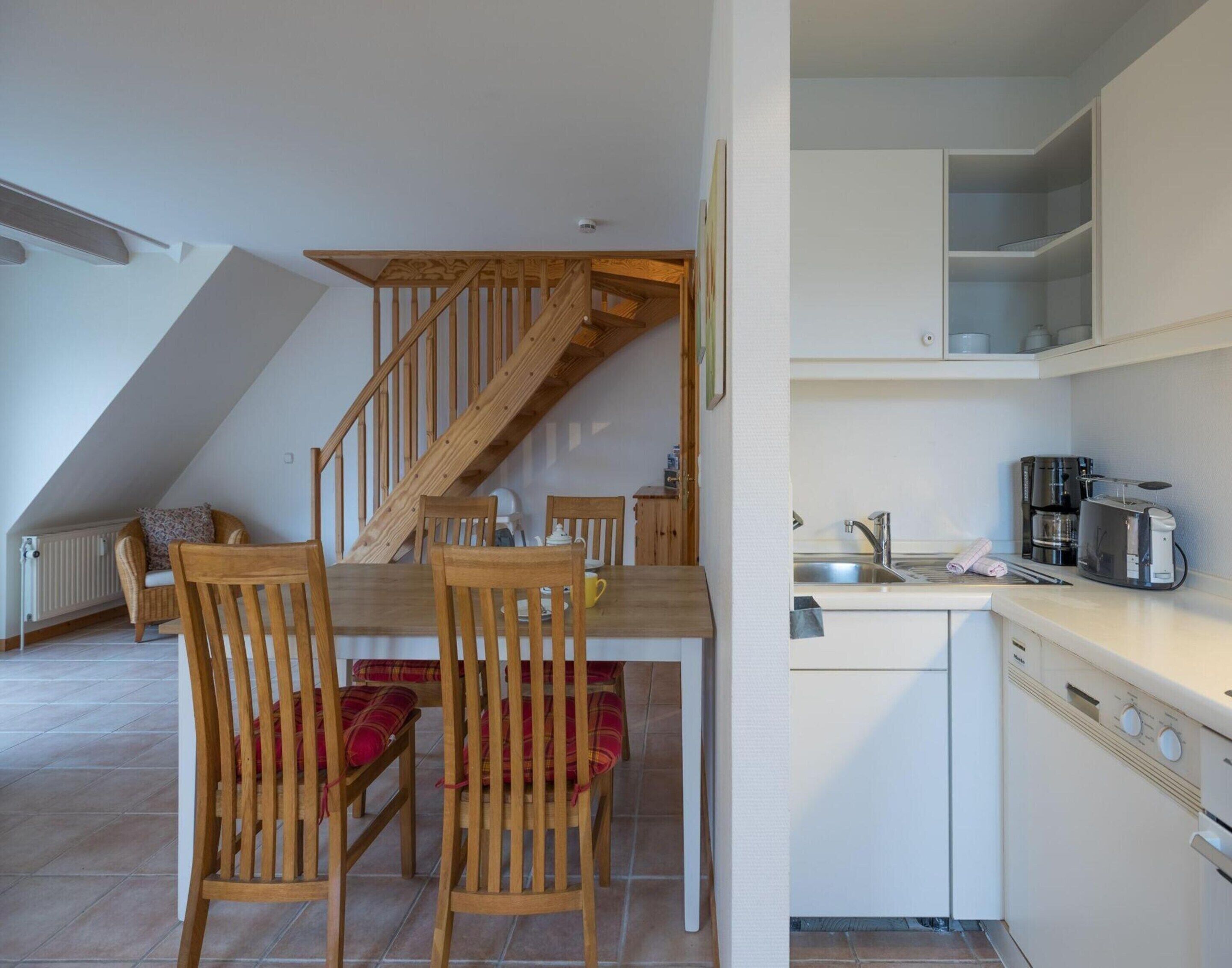 A dining area with wooden chairs and a table, a kitchen with white cabinets and appliances, and a wooden staircase.