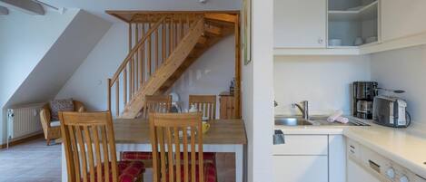 A dining area with wooden chairs and a table, a kitchen with white cabinets and appliances, and a wooden staircase.