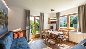 Een moderne woonkamer met een blauwe bank, een eettafel en stoelen. Er is een groot raam met gordijnen, een plafondlamp en uitzicht op groen buiten.