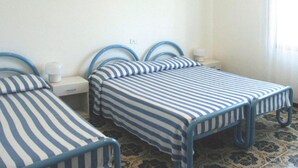 Una stanza con due letti, ciascuno dotato di una struttura in metallo blu e biancheria a righe. È presente un comodino e una lampada, con una presa elettrica sulla parete.