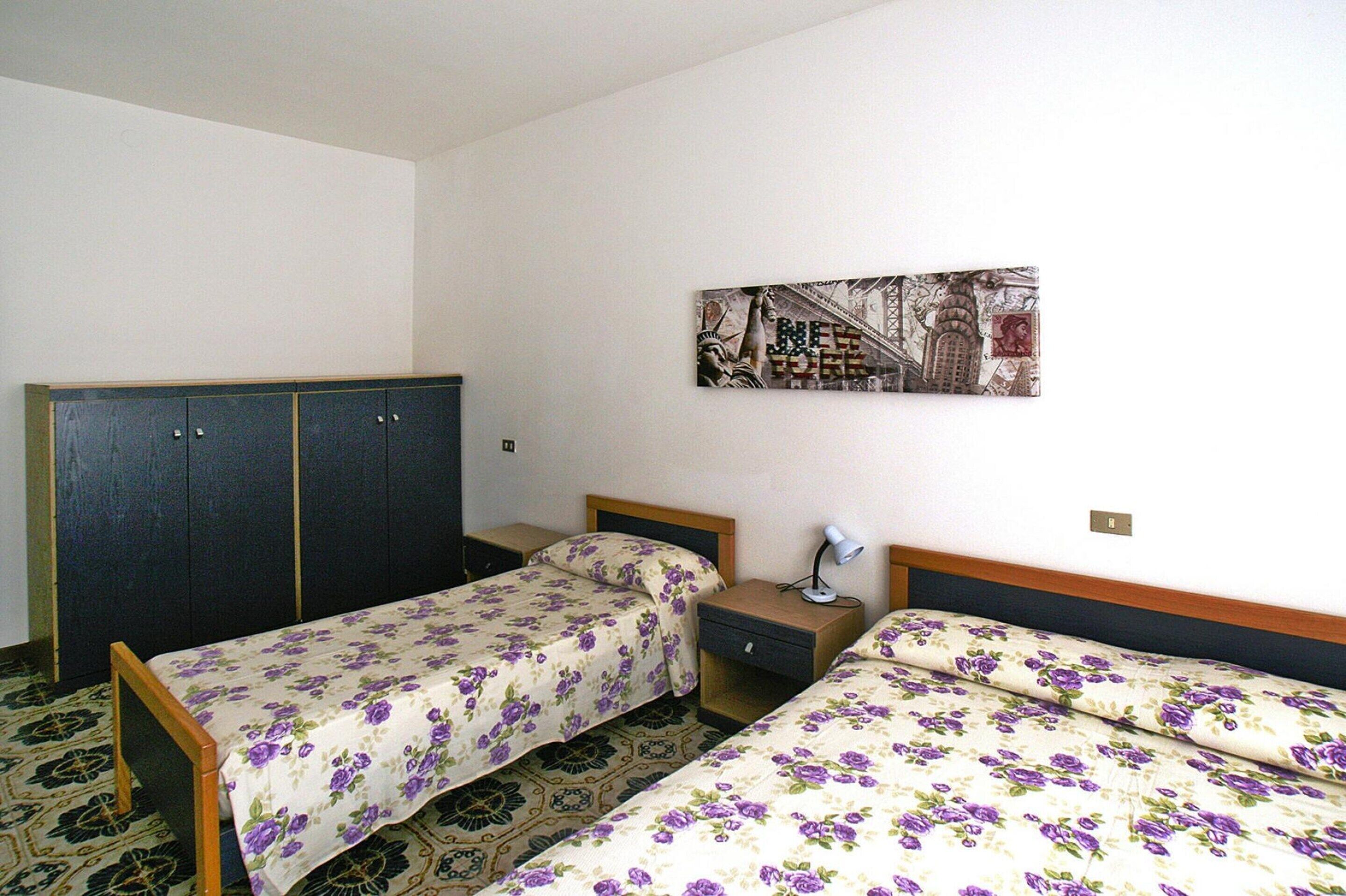 A hotel room with two single beds, a wardrobe, a nightstand, and a wall-mounted artwork.